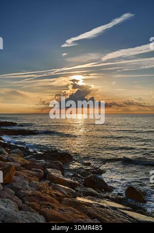 Vertikaler Blick auf einen dramatischen Sonnenaufgang über dem Mittelmeer, mit einer felsigen Küste und Sonnenstrahlen durch Cumulus Wolken in Alcossebre, Spanien Stockfoto