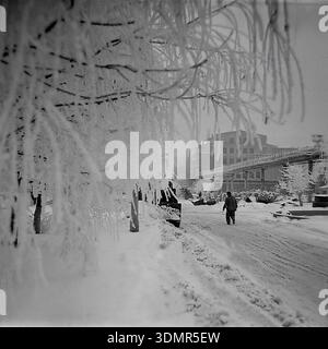Slawjansk, Ukrainische SSR, 1960er Jahre Ein einsamer Arbeiter läuft auf einer schneebedeckten Straße auf dem Gebiet des AIZ-Werks (Armature-Isolator Plant). Die Winterszene ist wunderschön eingerahmt von Baumzweigen schwer mit dickem weißem Raureif auf der linken Seite. Im nebeligen Hintergrund sind massive Industriegebäude und ein technologisches Rohrleitungsnetz über dem Kopf durch den Nebel sichtbar. Die Atmosphäre fängt die ruhige Entschlossenheit der Arbeit an einem kalten Wintertag in der industriellen Region Donezk ein. Ein authentischer Einblick in das tägliche Leben einer sowjetischen Fabrik im friedlichen Donbass. Stockfoto