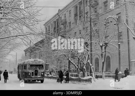 Slawjansk, Ukrainische SSR, 1960er Jahre Eine lebhafte Winterszene in der Schewtschenko Straße. Ein klassischer sowjetischer ZIS-155-Bus fährt auf der schneebedeckten Straße. Rechts steht ein prächtiges Wohngebäude aus der stalinistischen Zeit mit großen Bogenfenstern im Erdgeschoss und Balkonen in den oberen Stockwerken. Fußgänger laufen auf den verschneiten Gehwegen, die von frostbedeckten Bäumen gesäumt sind. Ein atmosphärisches Archivfoto, das öffentliche Verkehrsmittel und städtische Architektur in der friedlichen Region Donezk während der Sowjetzeit erfasst. Stockfoto