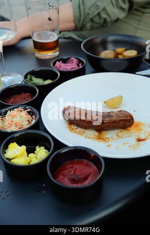 Nahaufnahme einer gegrillten Wurst auf einem weißen Teller, begleitet von kleinen Schüsseln mit verschiedenen Beilagen und Gewürzen einschließlich Gurken, Zwiebeln, Coll Stockfoto