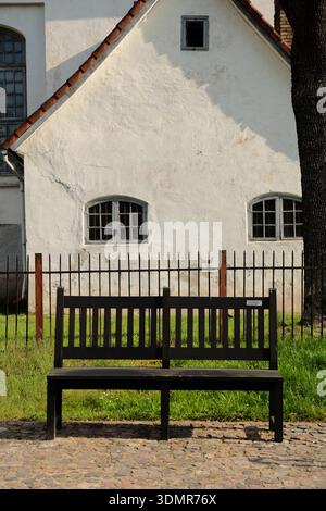 Minimalistische Szene mit einer schwarzen Holzbank auf einem Kopfsteinpflasterweg vor einem historischen weißen Putzhaus mit kleinen Fenstern. Weiche Sonnenliege Stockfoto