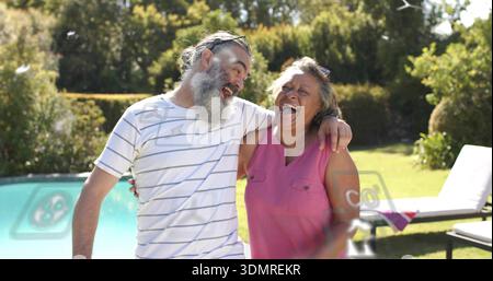 Lachendes Paar, das sich am Pool im Garten lehnt, Mann in gestreiftem Hemd, Sonnenbrille, Frau in rosa Tanktop Stockfoto