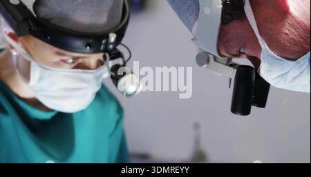 Zwei Chirurgen zur Durchführung eines Eingriffs in ODER, in blaublauem Peeling, Kappen, Masken, Scheinwerfern mit Lupen Stockfoto