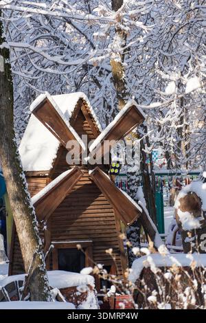 Rustikale Windmühle aus Holz mit schneebedeckten Blättern steht in einem verschneiten Wald, umgeben von hohen Bäumen und farbenfrohen Parkelementen Stockfoto