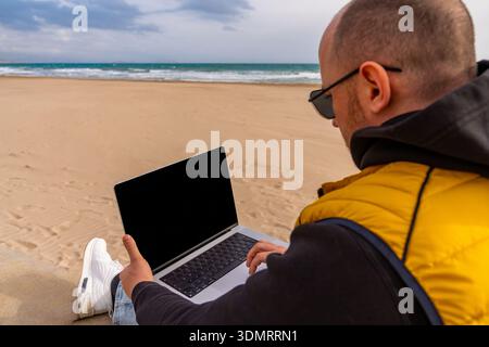 Mann im Hoodie, der ferngesteuert in der Nähe des Wassers arbeitet. Lässig gekleideter Programmierer, der sich auf den Bildschirm am Meer mit bewölktem Himmel konzentriert Stockfoto