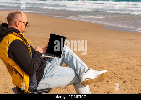 Person in Freizeitkleidung, die am Meer arbeitet. Gelegenheitsarbeiter in Jeans und Sonnenbrillen konzentrieren sich auf das Tippen in der Nähe von Wellen und Sandstrand Stockfoto