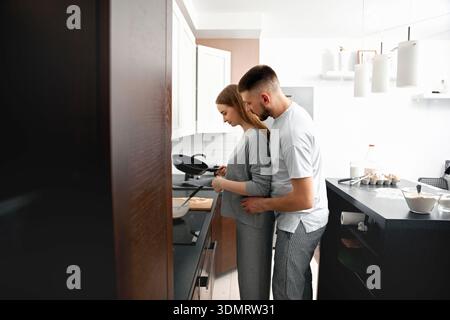 Ein Mann und eine Frau kochen an einem Wochenende in ihrer Küche. Die Frau steht am Herd, während der Mann hinter ihr steht und Nähe zeigt Stockfoto