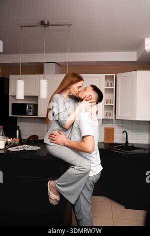 Ein Mann und eine Frau umarmen und küssen sich in einer Küche. Die Frau sitzt auf der Theke, und der Mann steht nahe. Die Umgebung ist gemütlich und zwanglos Stockfoto