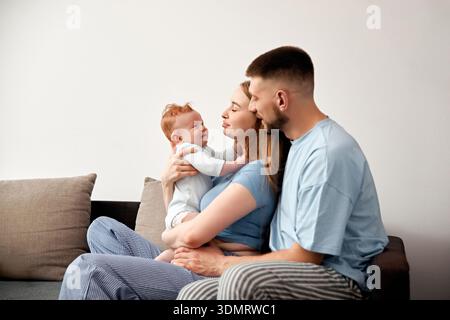 Eine Mutter und ein Vater sitzen mit ihrem Baby im Wohnzimmer auf einer Couch. Die Mutter hält das Baby nahe, während der Vater beide beobachtet und einen bo erschafft Stockfoto