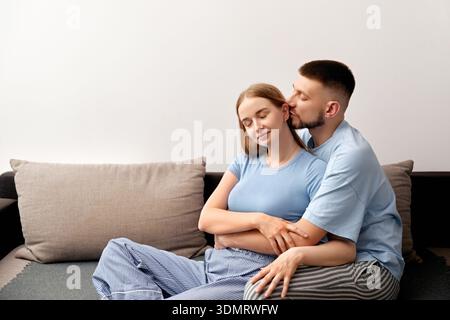 Ein junges Paar sitzt zu Hause dicht auf einer Couch. Der Mann beugt sich vor, um die Frau auf den Kopf zu küssen. Sie teilen ein Lächeln und genießen die Anwesenheit der anderen. Stockfoto