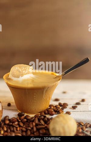 Nahaufnahme eines Eisglases mit schmelzendem Eiswürfel und Löffel. Kaffeebohnen auf dem Tisch verstreut. Stockfoto