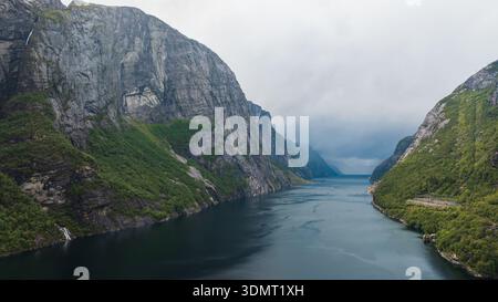 Aus der Vogelperspektive auf Lysefjord in Norwegen, mit steilen Klippen mit Grün, einem kleinen Wasserfall und tiefblauem Wasser unter einem bewölkten Himmel. Stockfoto