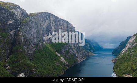Dramatische Luftaufnahme von Lysefjord in Norwegen mit steilen Klippen, einem kleinen Wasserfall, tiefblauem Wasser und einem nebeligen Horizont unter bewölktem Himmel. Stockfoto
