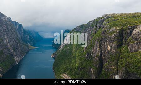 Blick aus der Vogelperspektive auf Lysefjord in Norwegen, mit zerklüfteten Klippen mit Grün, einem tiefblauen Fjord, Nebel, niedrigen Wolken und einer schmalen Straße entlang der Basis. Stockfoto