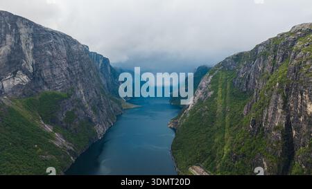Aus der Vogelperspektive des Lysefjords in Norwegen, mit steilen Klippen mit Grün, tiefblauem Wasser und niedrigen hängenden Wolken, die eine nebelige Szene schaffen. Stockfoto
