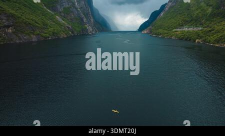 Aus der Vogelperspektive auf Lysefjord in Norwegen, mit ruhigem, dunklem Wasser, steilen grünen Klippen, einem gelben Kajak und einem kleinen Gebäude entlang der Klippe. Stockfoto