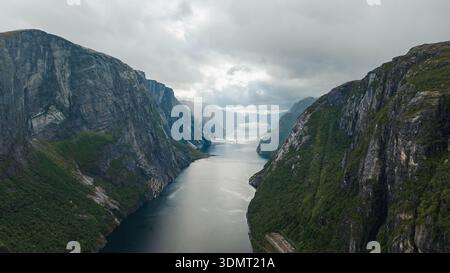 Dramatischer Blick auf Lysefjord in Norwegen mit steilen Klippen, zerklüfteten Felswänden, ruhigem, reflektierendem Wasser und einem bewölkten Himmel, der Atmosphäre verleiht. Stockfoto