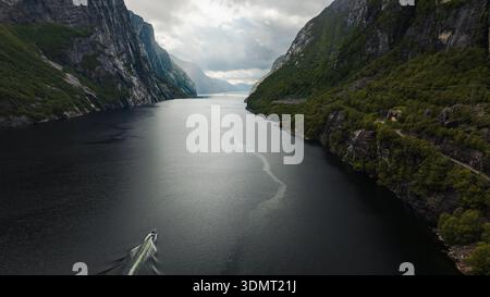 Blick aus der Vogelperspektive auf Lysefjord in Norwegen, mit felsigen Klippen mit Grün, einem Boot, das auf ruhigem Wasser eine Spur hinterlässt, und einer gewundenen Straße auf den rechten Klippen Stockfoto