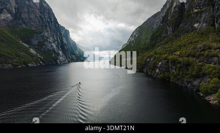 Blick aus der Vogelperspektive auf Lysefjord in Norwegen, mit steilen Klippen mit Grün, ruhigem, dunklem Wasser und einem kleinen Boot, das unter einem bewölkten Himmel wackelt. Stockfoto