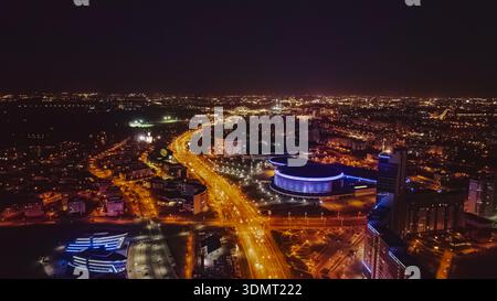 Nächtlicher Blick auf Minsk, Weißrussland, mit einem runden Gebäude mit blauen Lichtern, sich kreuzenden Straßen und modernen beleuchteten Gebäuden. Stockfoto
