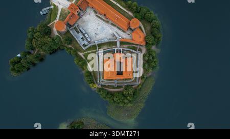 Blick von oben auf das Schloss der Insel Trakai mit roten, orangen Dächern, umgeben von ruhigem, blauem Wasser, üppigem Grün, Schilf und Bäumen in Litauen. Stockfoto