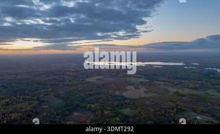 Ruhiger Blick aus der Luft auf einen großen reflektierenden See in Litauen, umgeben von Wäldern, Feldern und Ackerland mit herbstlichen Farben unter einem teilweise bewölkten Himmel. Stockfoto