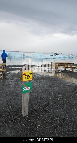 Ein Gletschergebiet in Island mit großen Eisbergen, einem hölzernen Pfosten mit Warnschildern, einer Person in einer blauen Jacke und einem felsigen Vordergrund unter einem bewölkten s Stockfoto