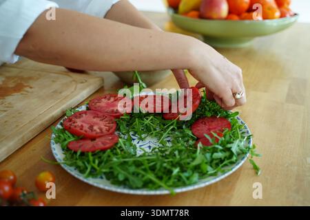 Frische Tomatenscheiben und Rucola sind auf einem dekorativen Teller angeordnet und zeigen eine lebendige Salatzubereitung. Eine Hand schneidet eine Tomate auf einem Holzbrett, Stockfoto