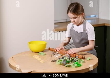 Mädchen rollt Teig mit Holznuten in der modernen Küche aus, schneidet Keksschneider aus und legt sie in der Küche auf Backpapier Stockfoto