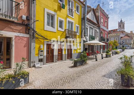 Lebhafte gelbe und rote Fassaden entlang einer schmalen portugiesischen Straße in Setubal, mit Cafés im Freien, Kopfsteinpflaster und einem Kirchturm in der Nähe Stockfoto