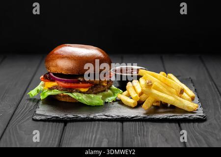 Rindfleisch-Cheeseburger mit Salat, Tomaten und Zwiebeln, serviert mit Pommes und Ketchup auf Schieferbrett Stockfoto