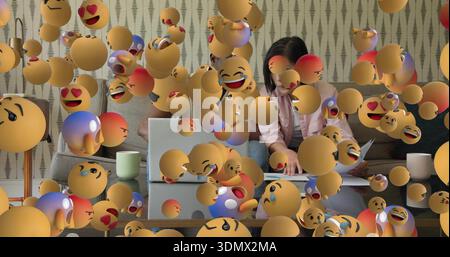 Arbeitende Koreanerin in hellrosa Hemd, die auf Laptop im Wohnzimmer schreibt, mit schwimmenden Emojis Stockfoto