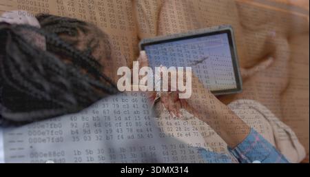Liegende Frau mit Zöpfen in hellblauem Hemd mit Eingabestift auf Tablette auf beigefarbenem Sofa, Daten-Overlay Stockfoto