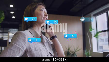 Sprechende asiatische Frau in beigefarbenem Hemd, Smartwatch, Telefon an Ohr im Büro, mit sozialen Alarmen Stockfoto