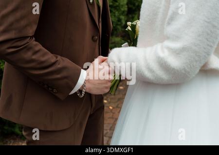 Ein zärtlicher Moment zwischen einer Braut und einem Bräutigam, der die Hände hält und ihre Hochzeitsoutfits mit dem weißen Pelzmantel und dem Blumenstrauß der Braut in einem üppigen G präsentiert Stockfoto