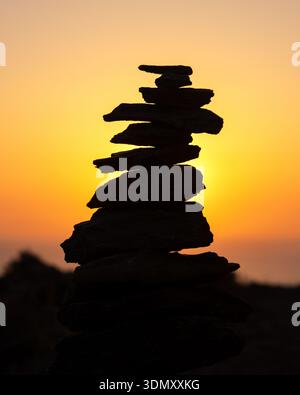 Silhouette aus ausgeglichenen Zen-Steinen, die bei goldener Stunde übereinander gestapelt sind, mit lebendigem orangefarbenem Abendhimmel und ruhigem Meer im Hintergrund, symbolisierend Stockfoto