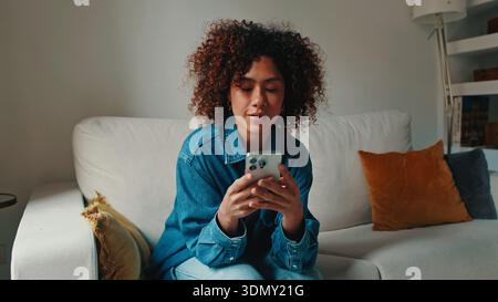 Eine Frau mit lockigen Haaren sitzt auf einem hellen Sofa in einem gemütlichen Wohnzimmer und konzentriert sich auf ihr Smartphone. Denim-Hemd, warmes orangefarbenes Kissen und weiche Beleuchtung Stockfoto