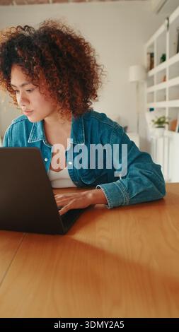 Eine Frau mit lockigem Haar und einer Jeansjacke sitzt an einem Holztisch und konzentriert sich auf ihren Laptop in einem hellen Zuhause. Ideal für Fernarbeit, Studium und e Stockfoto