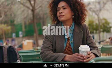 Eine stilvolle Frau mit lockigen Haaren sitzt in einem Café im Stadtpark und hält eine Einwegkaffee. Sie trägt einen braunen Mantel über einem gestreiften Hemd. Stockfoto