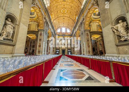 Im Inneren des Petersdoms in Vatikanstadt. Rotes Tuch säumt den Eingang des Kirchenschiffs mit Stühlen, Statuen, goldenen Decken und Kuppeln der Basilika St. Peter. Stockfoto