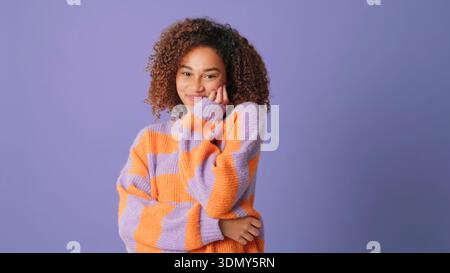 Ein fröhliches Porträt einer Frau mit lockigem Haar, die einen violett-orange gestreiften Strickpullover trägt, posierte vor einem weichen lila Hintergrund. Warm, freundlich Stockfoto