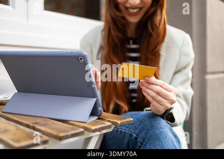 Eine lächelnde Frau, die draußen sitzt, benutzt ein Tablet zum Online-Shopping und hält eine gelbe Bankkarte in der Hand. Die entspannte Atmosphäre lässt eine nahtlose digitale transa entstehen Stockfoto