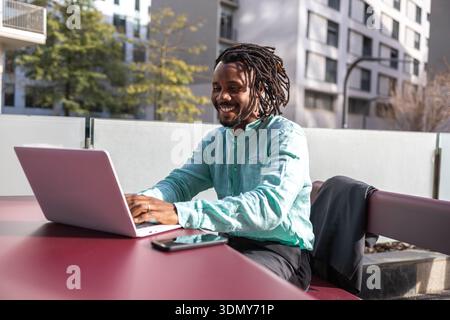 Afroamerikanischer Geschäftsmann mit Dreadlocks lächelt einen Laptop an, während er draußen arbeitet. Er sitzt an einem Tisch in einer urbanen Umgebung und genießt einen sonnigen Tag Stockfoto
