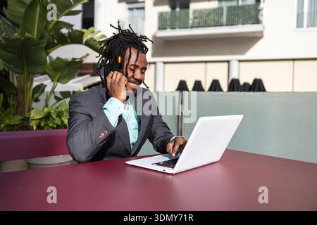 Afroamerikanischer Geschäftsmann mit Dreadlocks, der einen Anzug trägt und draußen an einem Tisch sitzt. Er ist mit einem Anruf beschäftigt und arbeitet an einem Laptop, lächelt und Stockfoto