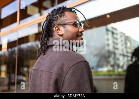 Afroamerikanischer Geschäftsmann mit Dreadlocks und Brille, der selbstbewusst draußen steht. Er trägt einen Mantel, der den modernen urbanen Geschäftsstil symbolisiert Stockfoto