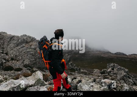 Ein Mann mit Outdoor-Ausrüstung und Rucksack steht auf felsigem Gelände mit Blick auf einen majestätischen Berg, der in Nebel gehüllt ist und den Geist des Abenteuers A verkörpert Stockfoto