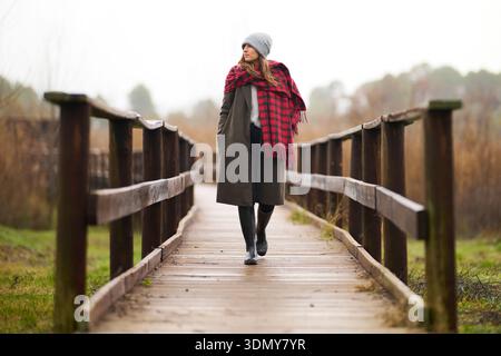 Eine blonde Frau in einem gemütlichen Mantel und einem roten Decken-Schal geht an einem regnerischen Tag auf einem Holzweg entlang. Sie trägt eine Beanie und Stiefel, die Komfort und e verkörpern Stockfoto