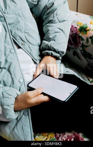 Eine Person, die eine hellblaue Steppjacke trägt, hält ein Smartphone mit einem leeren Bildschirm. Das Individuum sitzt auf einem Stuhl mit Blumenmuster, Creati Stockfoto