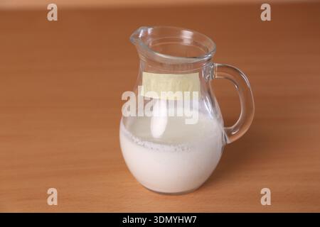 Ein Glaskrug mit frischer Milch steht auf einem Holztisch in einer Küche. Die Einstellung ist hell, und die Kanne hat ein einfaches Etikett. Es gibt kein anderes Essen Stockfoto