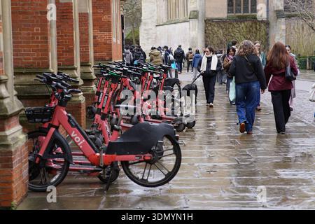 Eine Reihe roter VOI Elektroroller, die an einem nassen Wintertag vor dem Eingang des historischen Cambridge College geparkt sind und an dem Fußgänger vorbeikommen. Cambridge, England Stockfoto